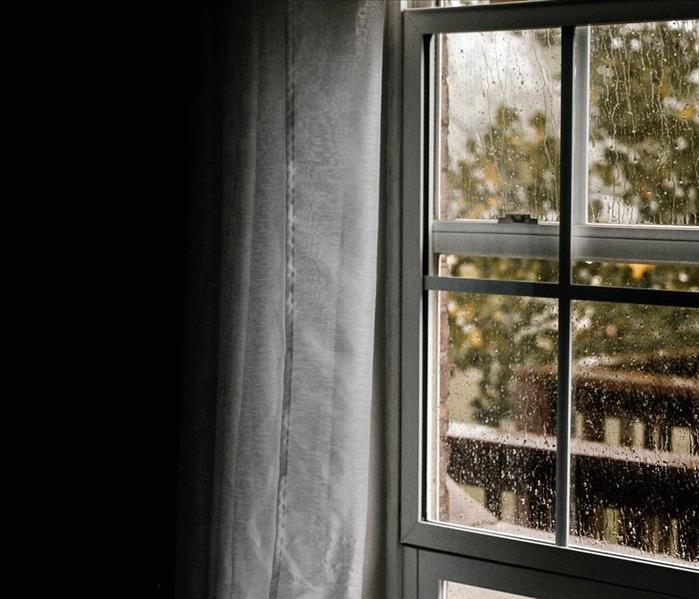 A window and curtain on a rainy day, covered in raindrops with the sunlight coming in.